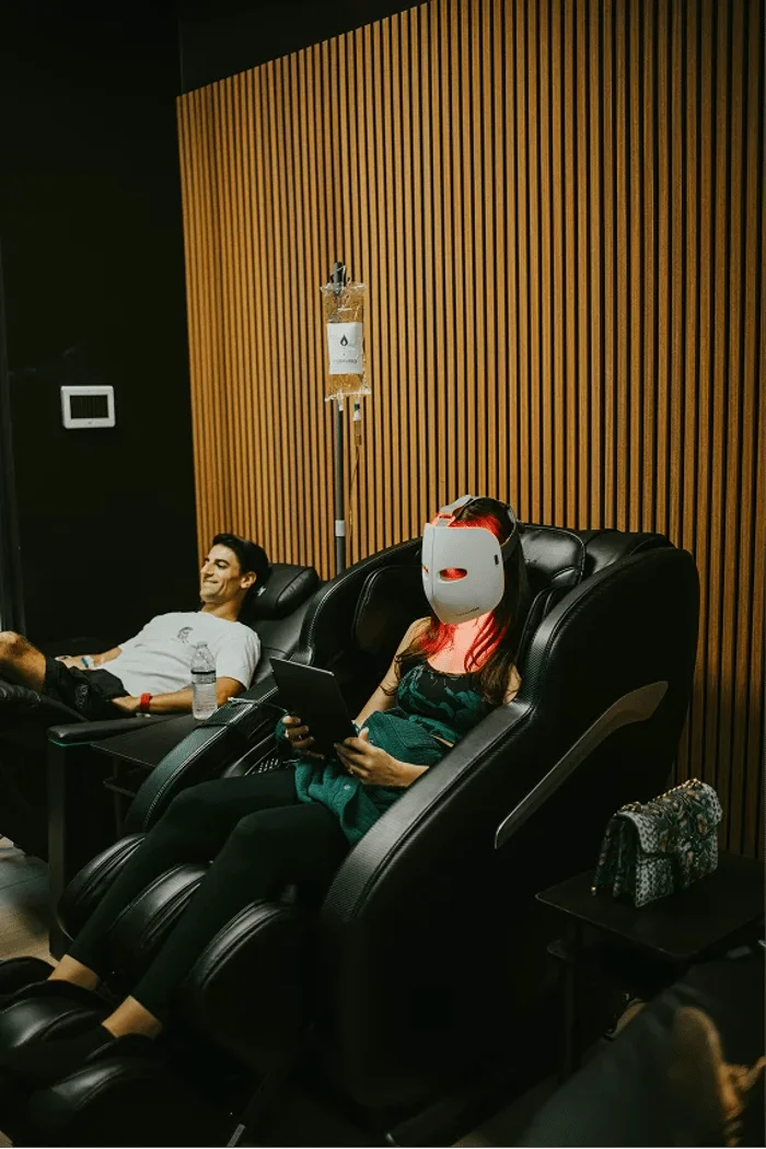 A woman in a face mask holds a tablet while receiving IV therapy