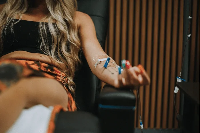 A person with long blonde hair receiving an IV infusion in their left arm.